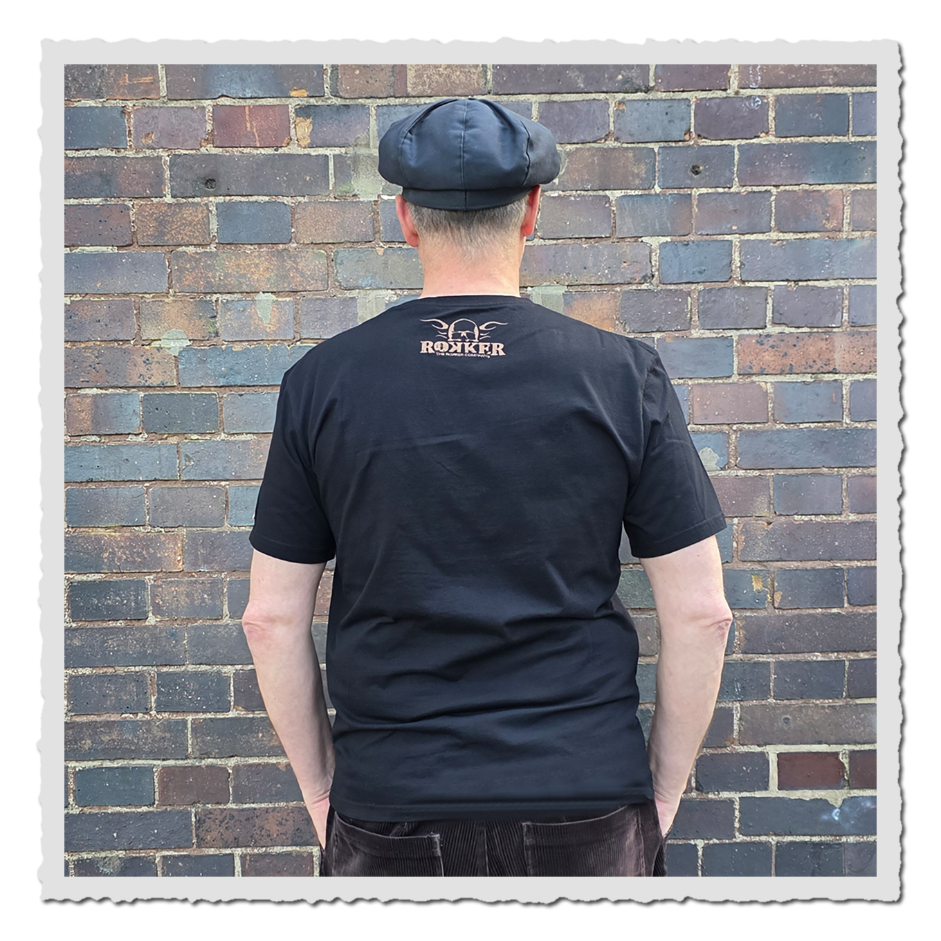 Mann von hinten in schwarzem T-Shirt mit kleinem Nackenprint, dunkler Kappe, vor mehrfarbiger Backsteinwand.